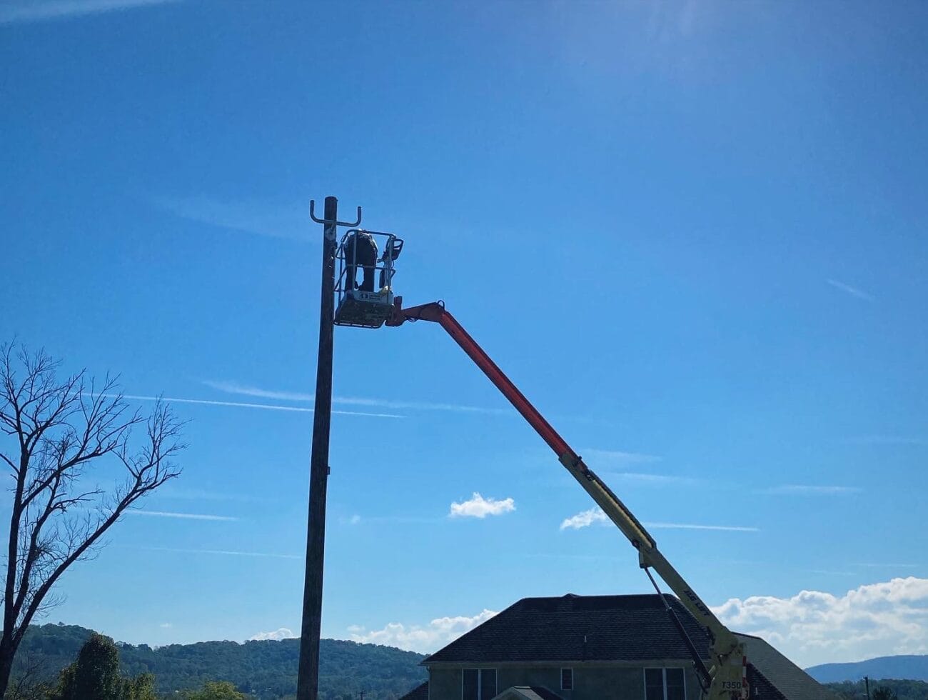 A worker in a bucket lift performs maintenance on a utility pole under a clear blue sky, with a house and hills in the background.