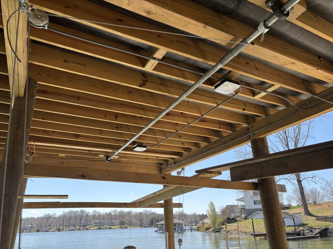View from under a wooden dock roof showing exposed beams and pipes, overlooking a lake with houses and trees in the background on a sunny day.