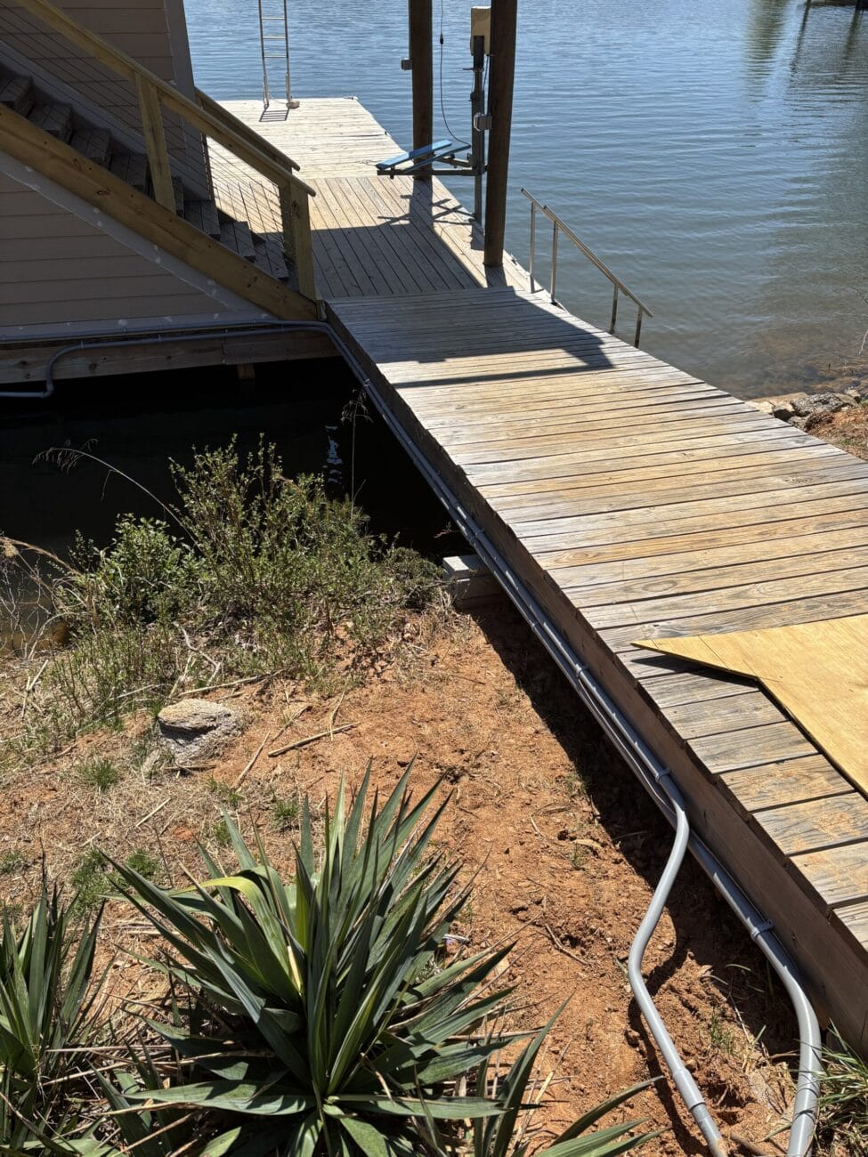 A wooden dock with stairs leads to a lakeside house; electrical conduit runs along the ground beside dry soil and green plants near the dock.
