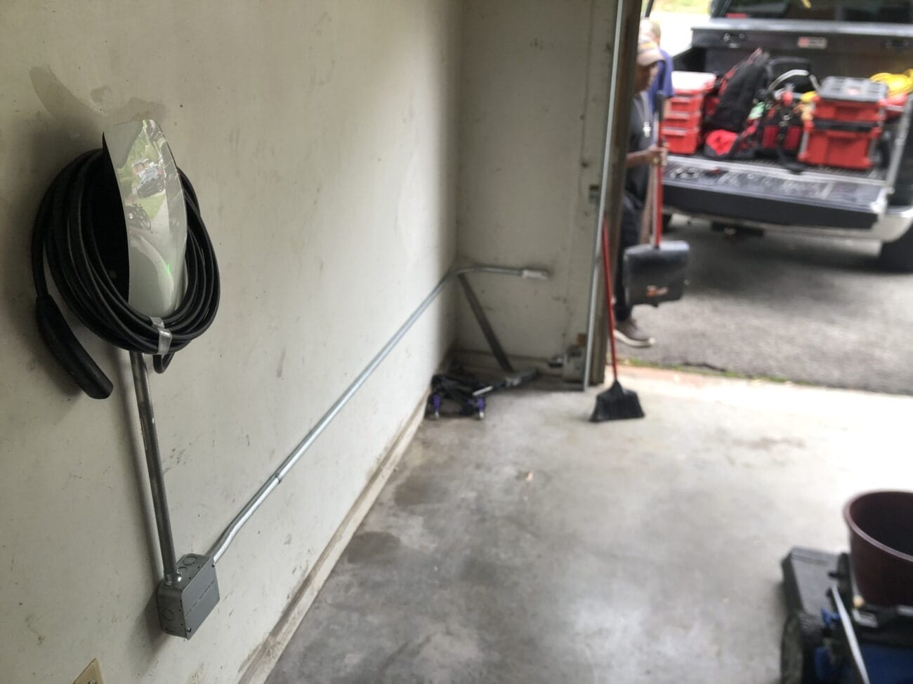 Electrical conduit runs along a garage wall, ending at an outlet box; a coiled cable hangs above. Tools and equipment are visible in a truck parked just outside the open garage door.