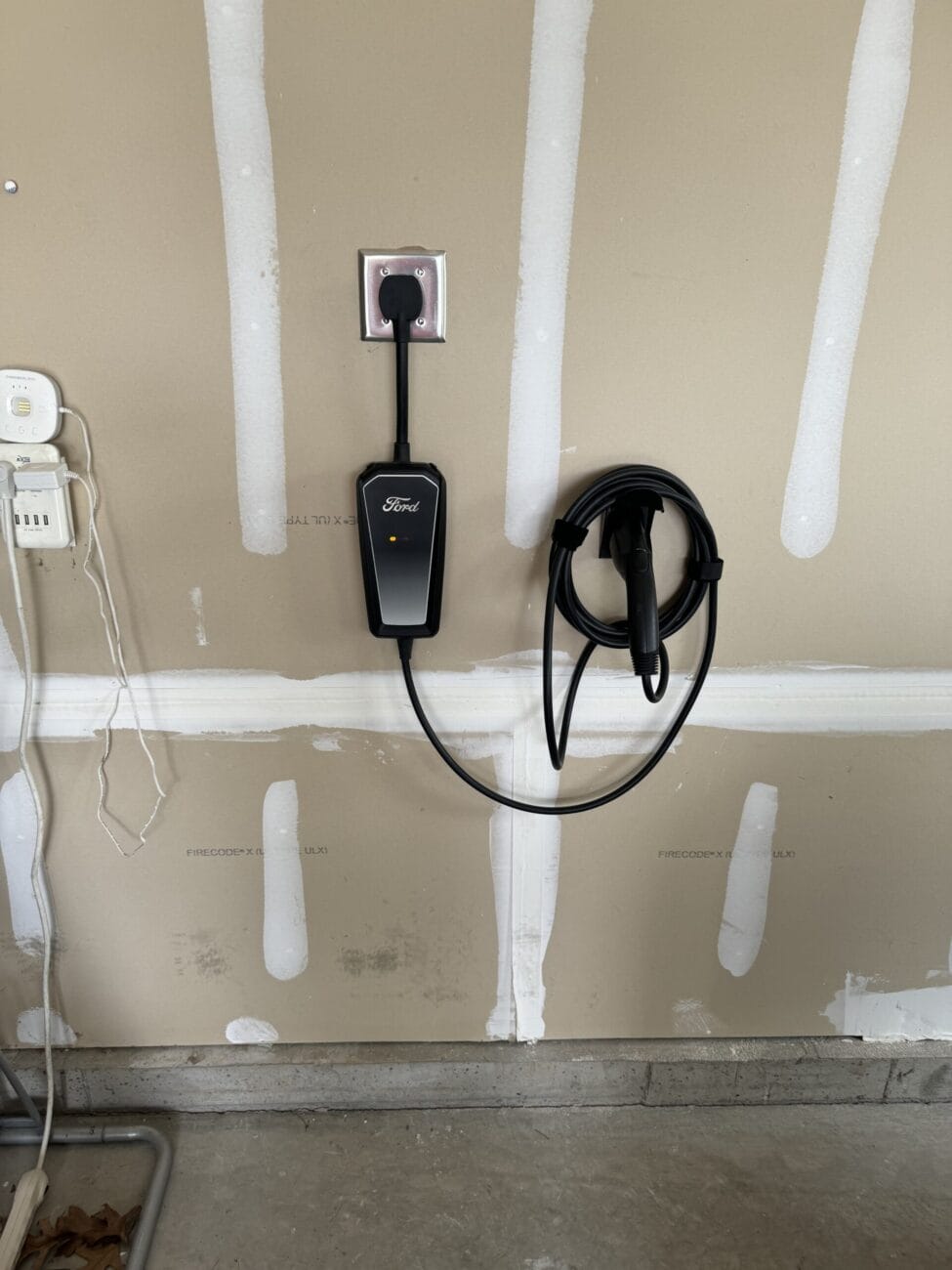 A Ford electric vehicle charger is plugged into a wall outlet in a garage, with the charging cable coiled neatly on a mounted hook.