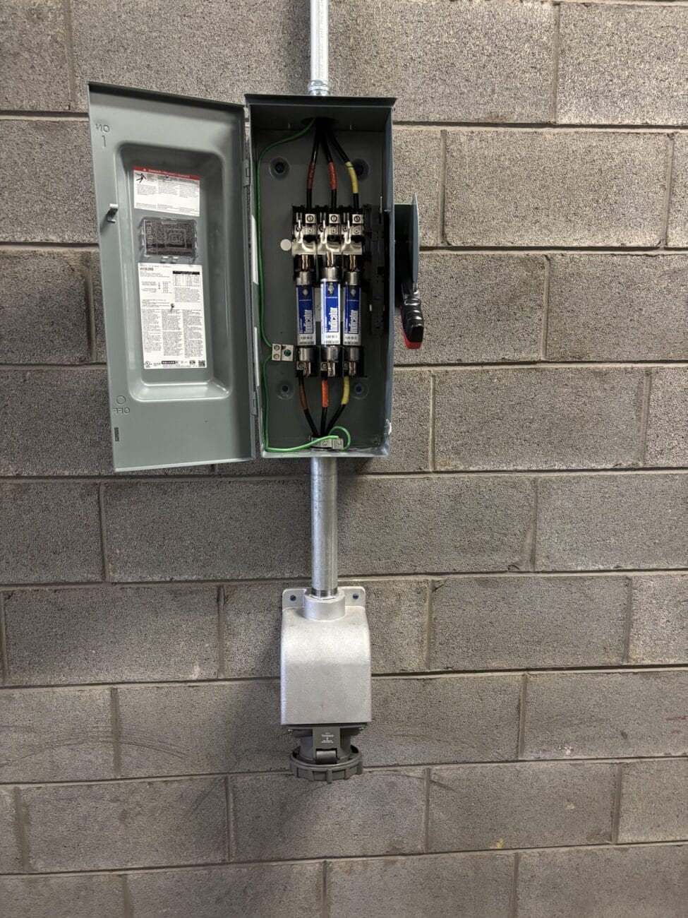 Electrical fuse box with open door mounted on a gray cinder block wall, showing wiring and fuses inside, with conduit and a power outlet below.
