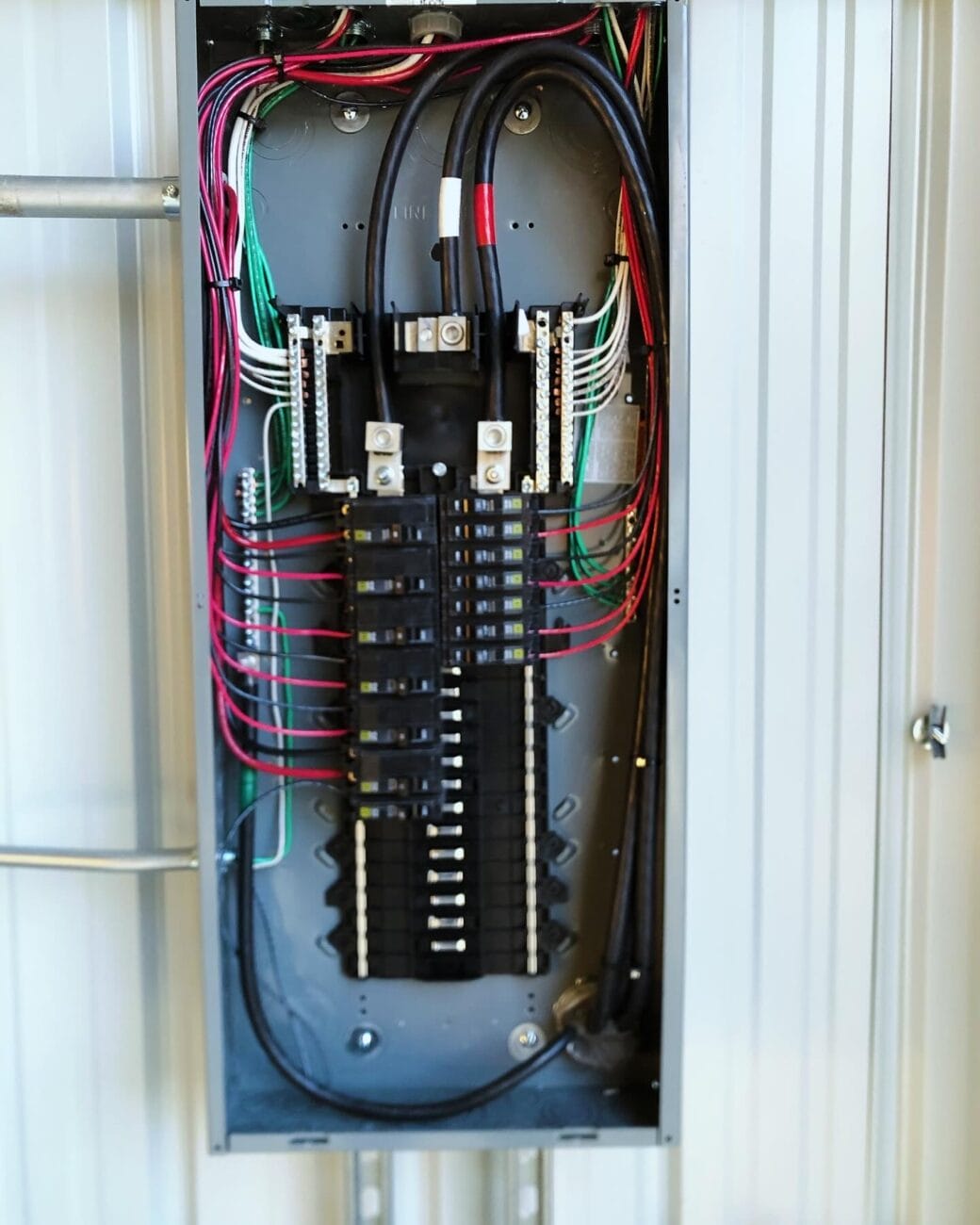 An open electrical breaker panel with multiple circuit breakers, black and red wires, and green grounding wires, mounted on a light-colored wall.