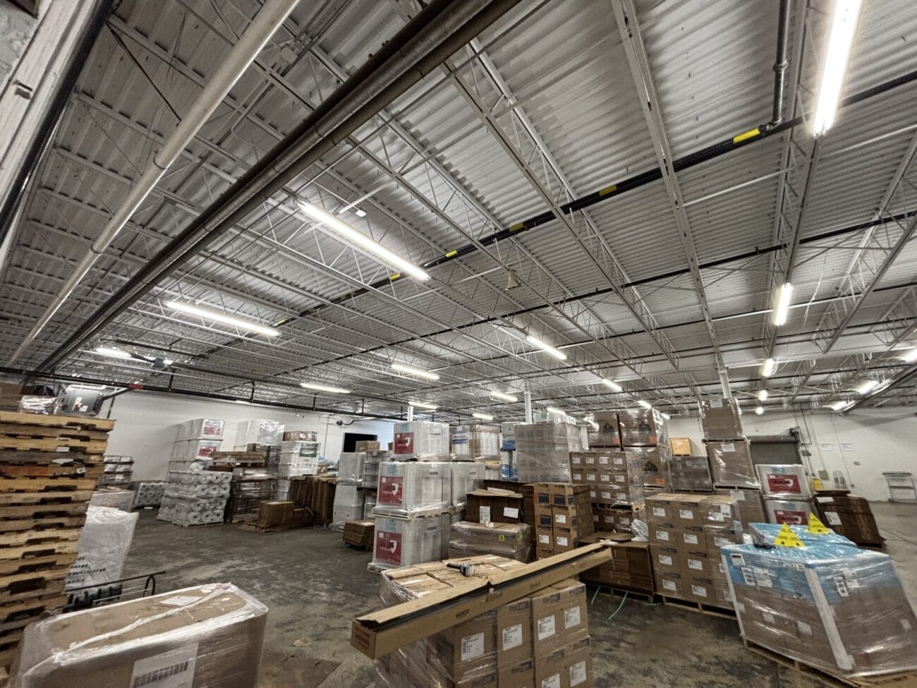 A warehouse interior with stacked pallets, boxes, and various supplies organized throughout the space under a metal ceiling with fluorescent lighting.