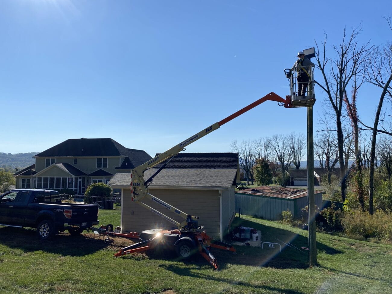 A worker in a bucket lift is repairing or installing equipment on a utility pole in a residential backyard on a clear day.