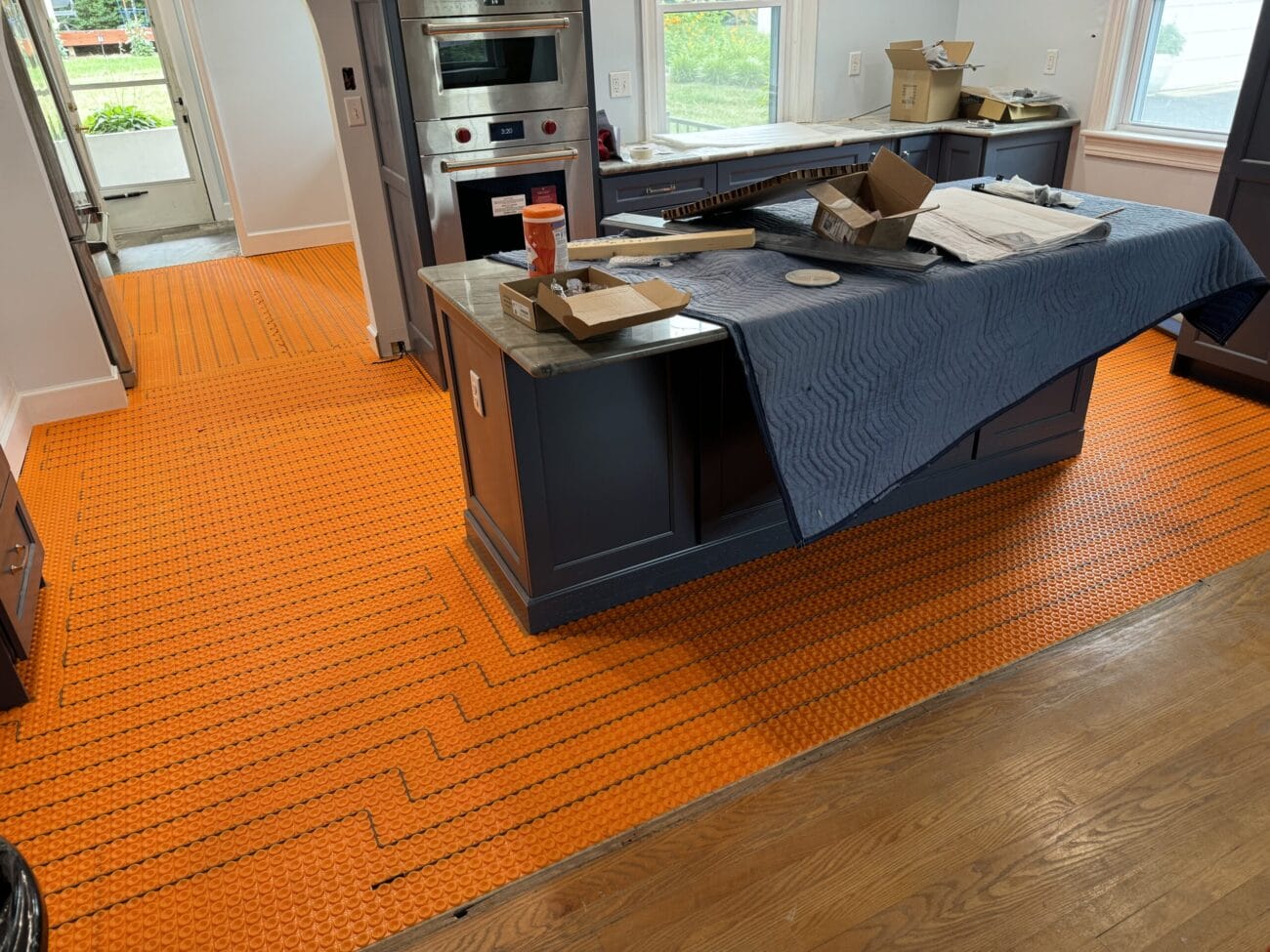 A kitchen under renovation with bright orange underfloor heating mats installed, a covered island, and construction materials scattered on counters.