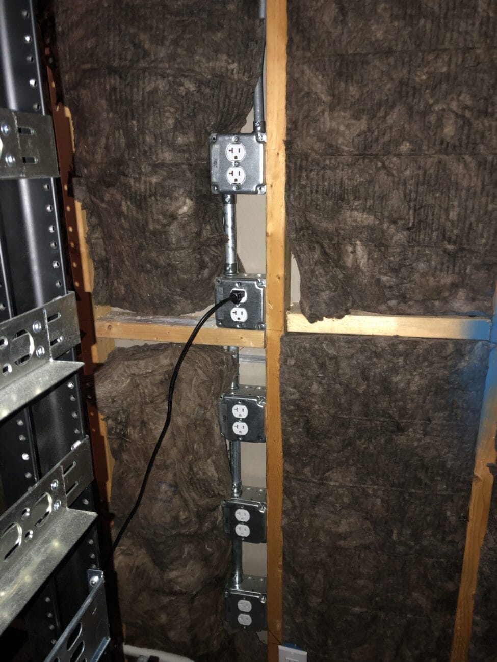 Metal electrical outlet boxes installed vertically on exposed studs with insulation in the wall behind them. A black power cord is plugged into one of the outlets.